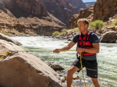 The Definitive 5:1 Rigging Guide for Whitewater Rafters A male rafter in a life vest managing a 5:1 rope rescue system on a rocky riverbank.
