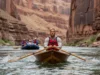Dory vs Raft: Grand Canyon Performance & Safety Analysis A skilled male guide rows a wooden dory through a Grand Canyon rapid, with an inflatable raft visible in the background for comparison.