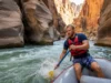 Undercut Rocks Rafting: Spotting & Avoiding River Hazards A male rafter in a life vest skillfully navigates a raft away from a dangerous undercut rock wall in a challenging whitewater river canyon.