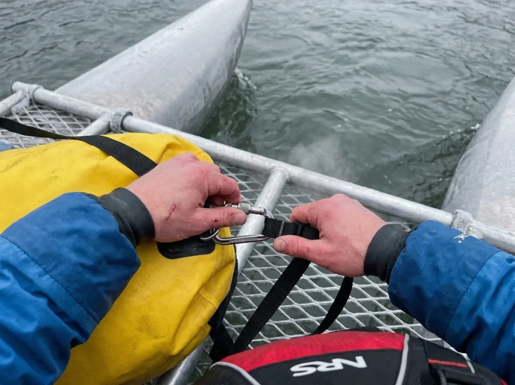 Hands clipping a dry bag to a cataraft frame with a carabiner above cold gray river water