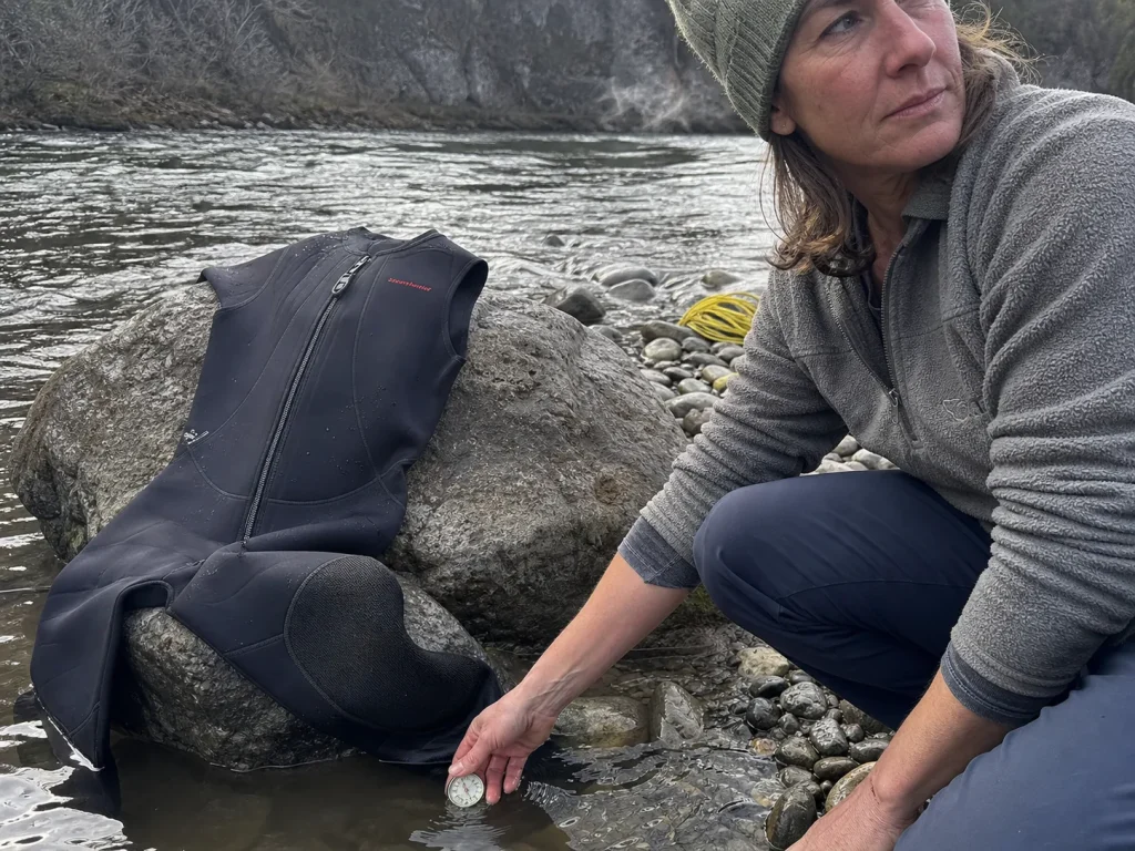 Rafter checking river water temperature against a hanging 3mm Farmer John wetsuit on a cold morning