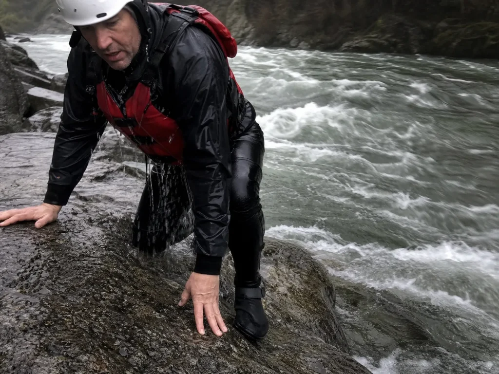Rafter climbing onto shoreline rocks after a cold swim with neoprene booties still on