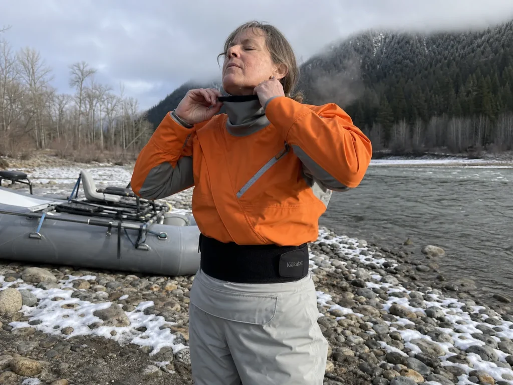Boater in a drysuit sealing the neck gasket beside a raft on a frigid snowmelt river in early season