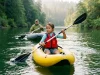 Kids Rafting Paddle vs Kayak Paddle What Yours Needs Young child paddling a kids rafting paddle in an inflatable kayak beside a family raft on a calm river