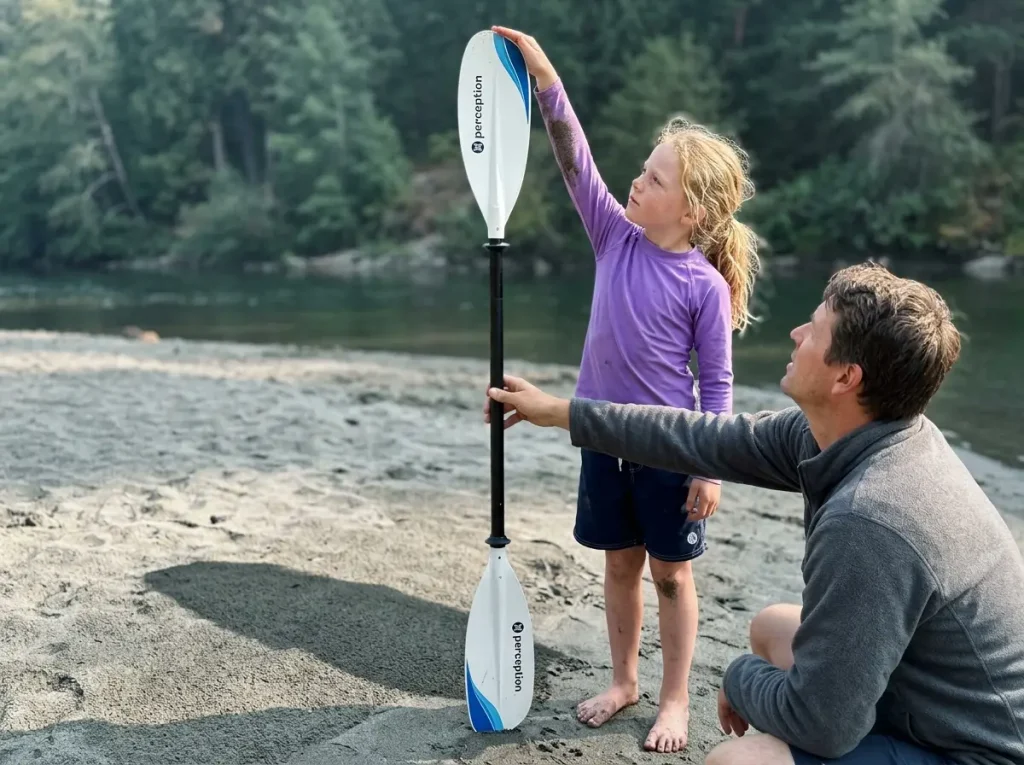 Parent holding a kids kayak paddle upright next to a standing child to check paddle length for correct sizing