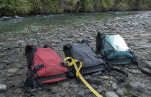 PFD Sizing Guide for Rafters Where Medium Isn’t Medium Three whitewater life jackets laid out on a gravel riverbank next to a soft measuring tape