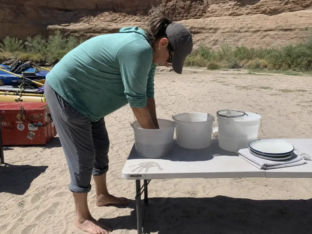 Woman bent over a low camp table washing dishes on a river sandbar, showing why table height matters