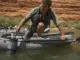 Drag Bags vs Soft Storage for River Camping Loaded mesh drag bag clipped to a raft D-ring, towing alongside the boat on a green desert river