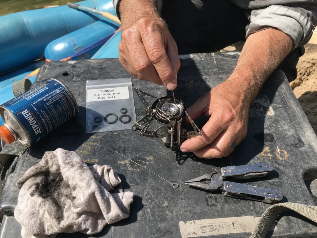 Backup camp stove with spare o-rings and a jet cleaning tool laid out at a river camp