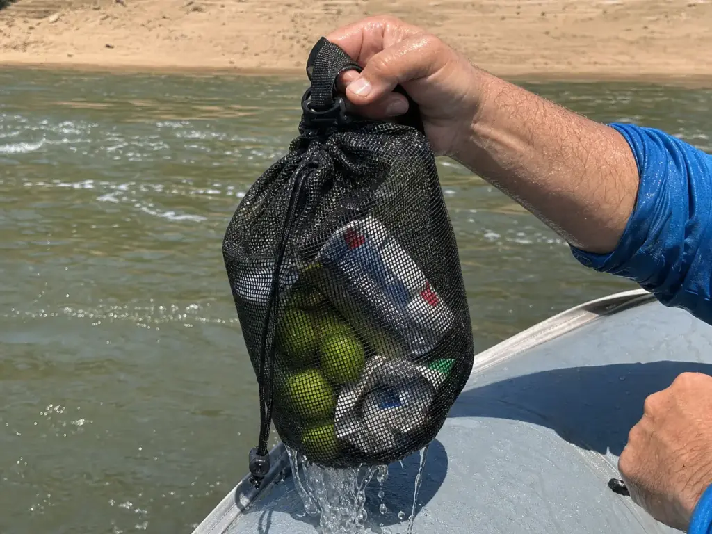 Hands pulling a dripping mesh drag bag out of the river, cans and produce visible through the wet mesh