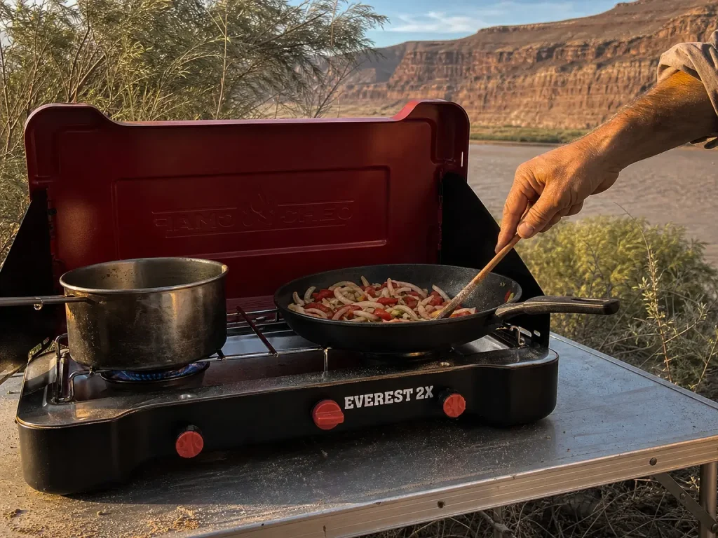 Two-burner camp stove with windscreen up cooking a group meal at a river camp