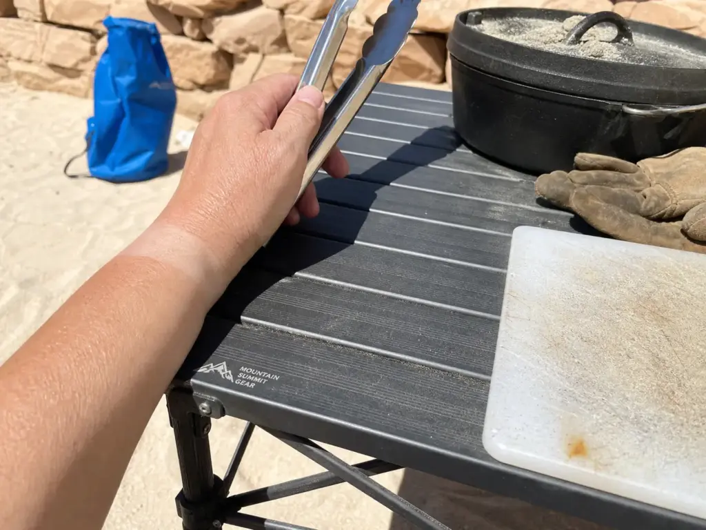 Hand reaching over a sun-baked aluminum camp table top with a Dutch oven, showing heat buildup