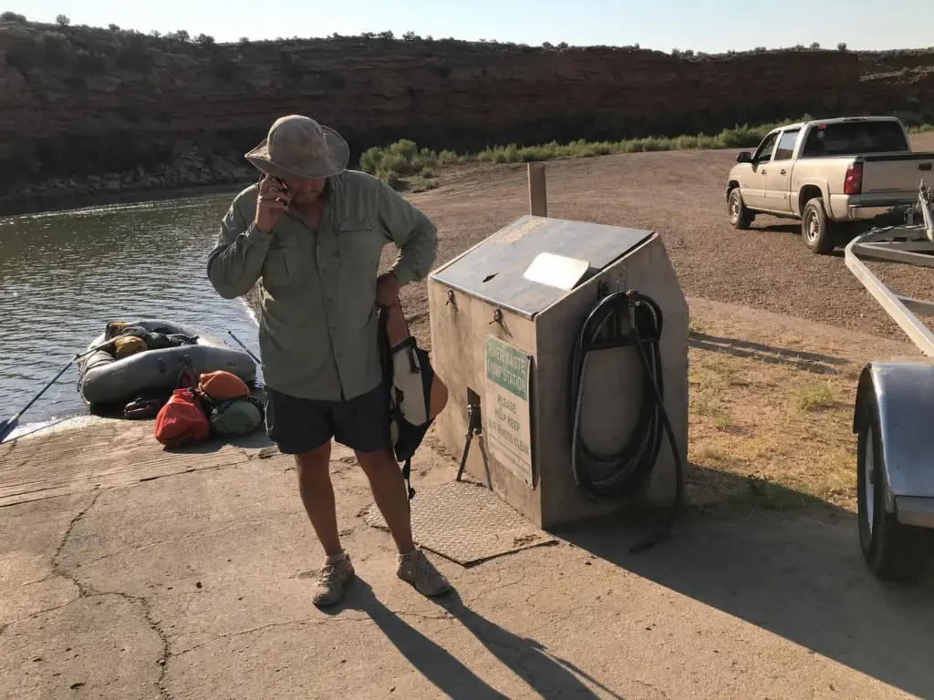 A boater on the phone beside a river takeout waste dump station, checking cleanout rules